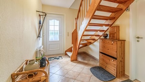 Eine Holztreppe führt zu einer zweiten Etage, eine Tür mit Fenster, ein kleiner Holzschrank und ein Korbwesen mit Gegenständen auf einem Fliesenboden.