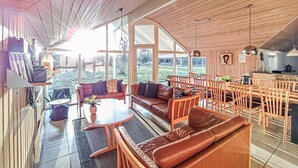 Amplia sala de estar con un sofá grande, mesa de comedor y sillas. Grandes ventanales que permiten la entrada de luz natural y vistas al exterior.