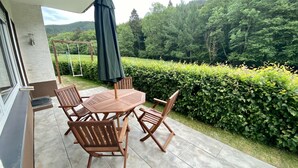 An outdoor patio with a wooden table and chairs, a green umbrella, and a view of a lush garden and forest. - beautiful, very quiet apartment with terrace  (Winterberg)