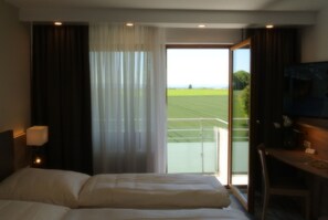 A hotel room with a bed, a desk, a chair, a lamp, and a view of a green field through a large window. - Studio apartment with a balcony and a small kitchen (Ostfildern)