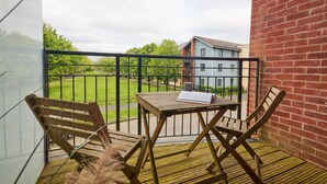 Un balcon avec une table en bois et deux chaises, un livre posé sur la table, et une vue sur un quartier résidentiel avec des maisons et un champ d’herbe.