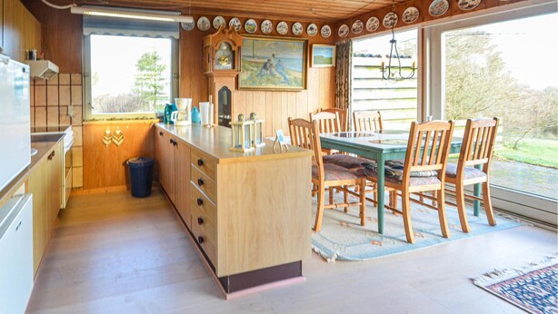 A kitchen with wooden cabinets, a dining table with chairs, a clock, and a painting on the wall.
