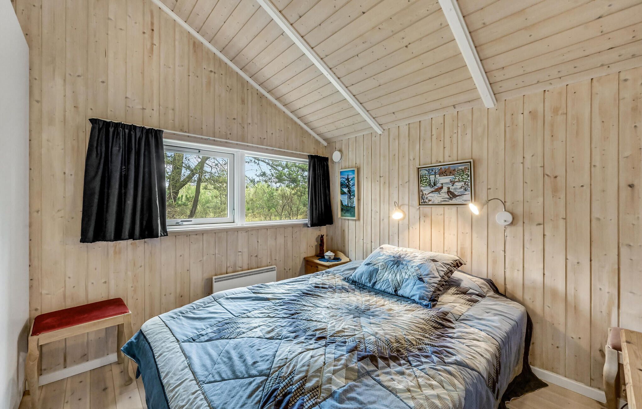 Une chambre avec des murs en bois, un grand lit recouvert d’une courtepointe, une fenêtre avec des rideaux, une table de chevet avec une lampe et un tableau accroché au mur.