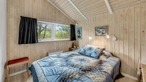 A bedroom with wooden walls, a large bed with a quilt, a window with curtains, a bedside table with a lamp, and a painting on the wall. - Beach front home in Hurup Thy with WiFi (Hurup Thy)