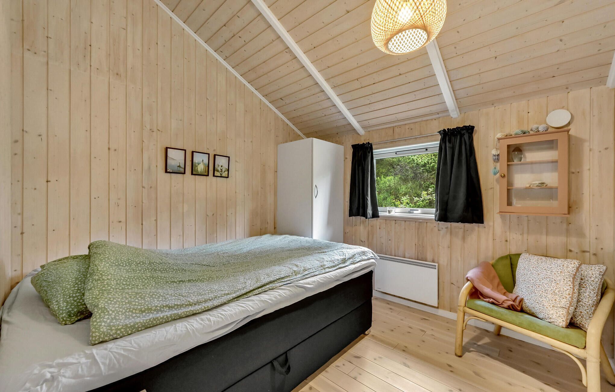 Ein Zimmer mit Holzwänden, einem Bett, einem Sessel, einem fenster mit Vorhängen und einem an der Wand befestigten Schrank.