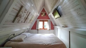 Un dormitori amb sostre inclinat, un llit amb roba de llit blanca, una taula de nit, una làmpada, una finestra amb cortines i un quadre al mur.
