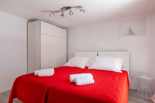 Una cama bien hecha con una colcha roja y almohadas blancas, un armario blanco y una mesita pequeña blanca.