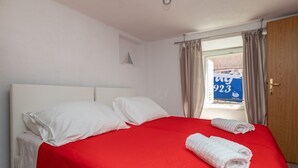 A neatly made bed with white pillows and a red bedspread, a rolled towel on the bed, and a window with curtains. - Old Town Charming Nest (Dubrovnik)