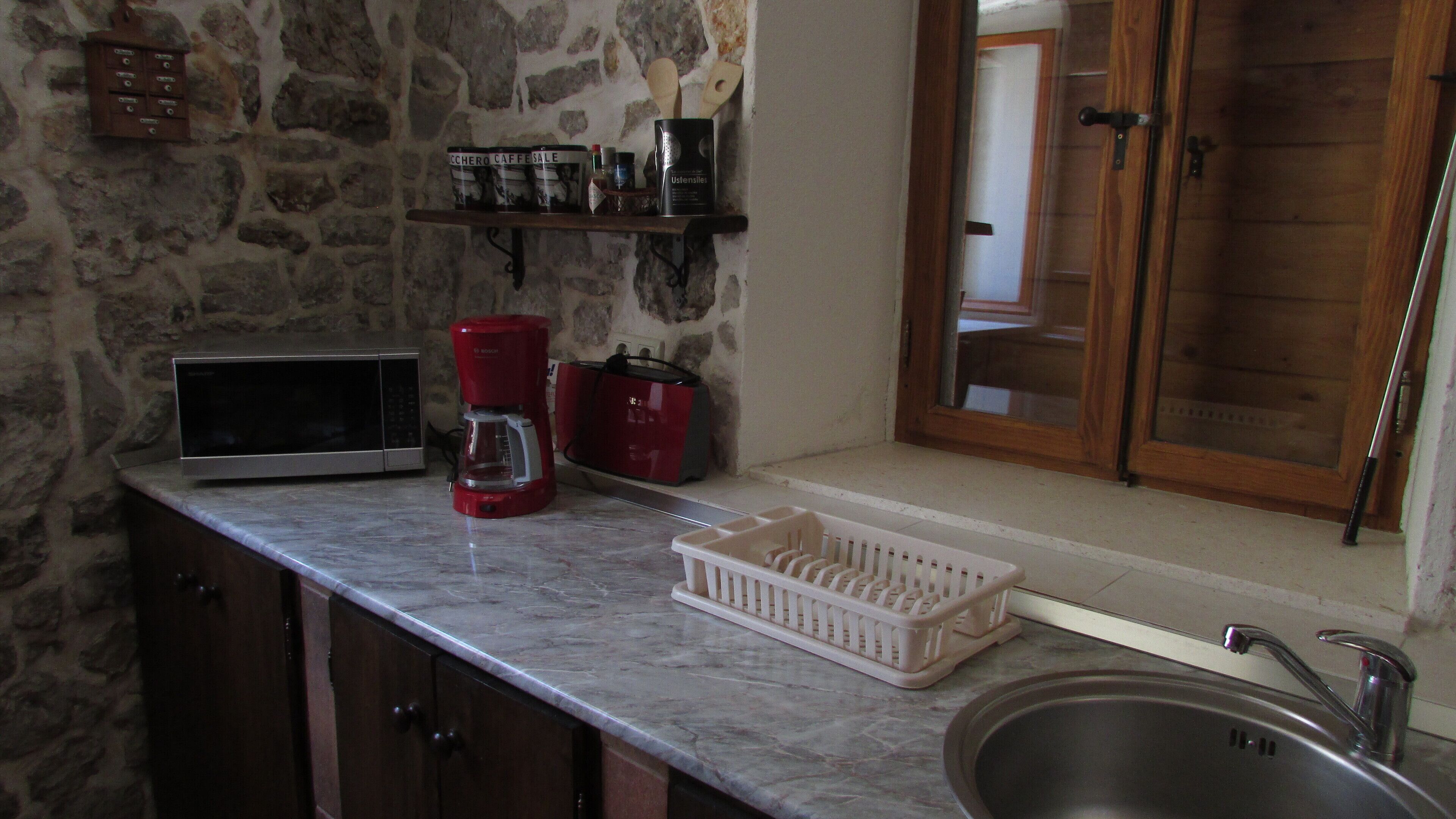 Una cocina con pared de piedra, microondas, cafetera roja, cesta y pileta.