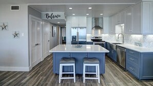 A modern kitchen with a central island, two bar stools, and a 'Believe' sign above the door. - Sea Gull #908: Beachfront 2 Bedroom 2 Bathroom With Spectacular Views and 24 Hour Management (Port Aransas)
