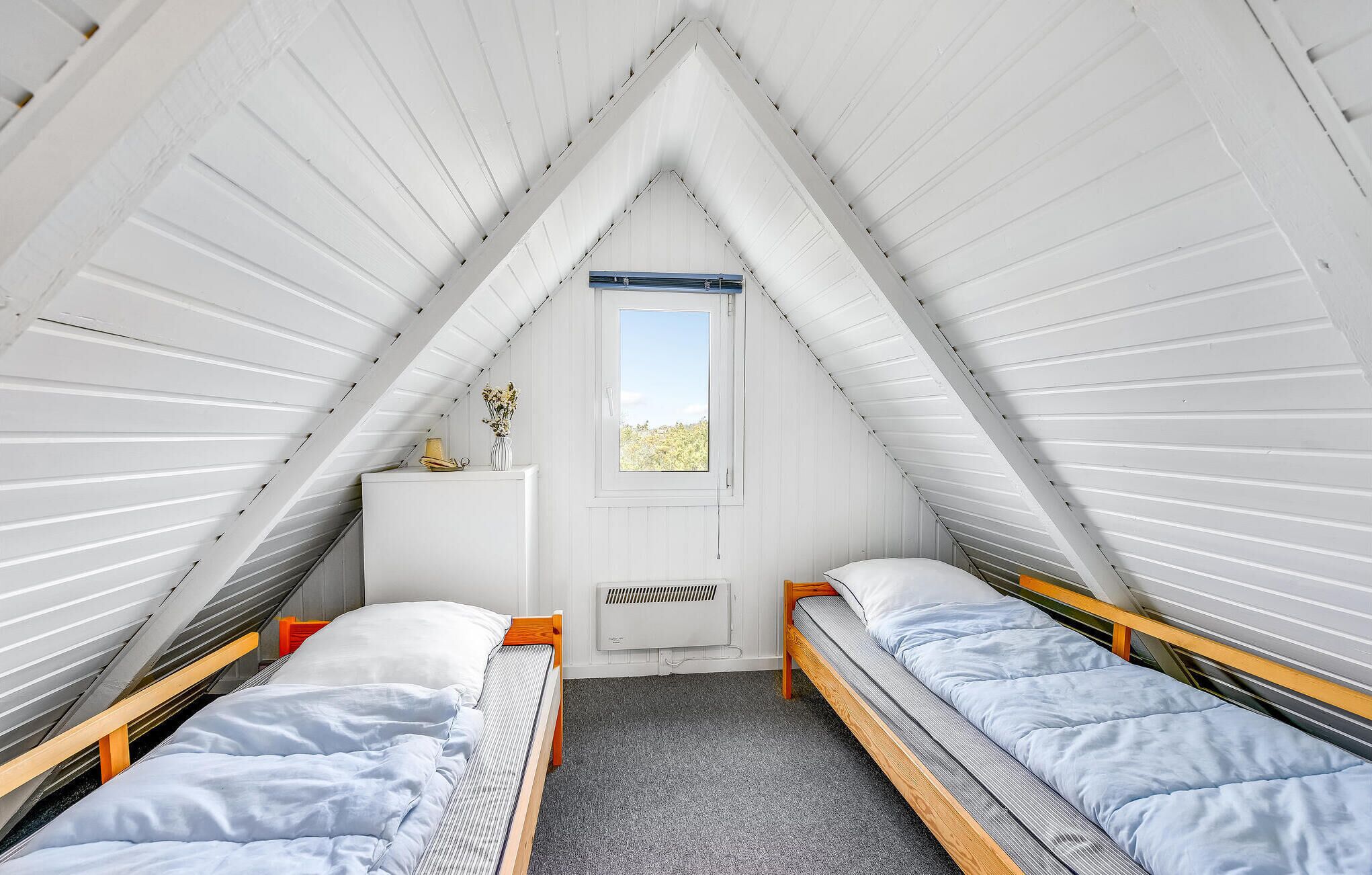 Ein kleines, weißes Schlafzimmer im Mansardenstil mit zwei Einzelbetten, einer weißen Kommode und einem Fenster mit Blick auf Bäume.