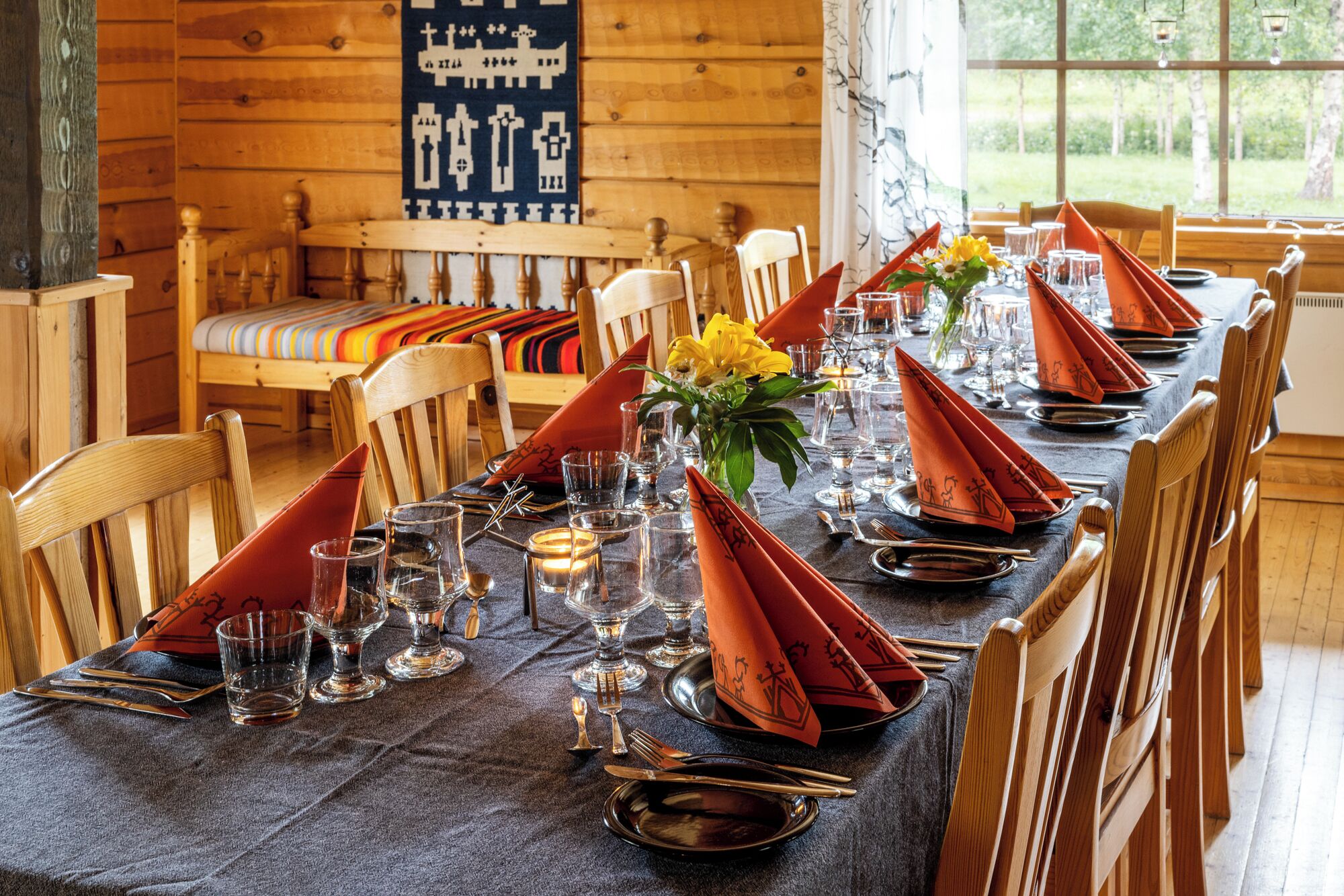 Een eettafel gedekt met oranje servetten, glazen en bestek, in een kamer met houten wanden en een raam.