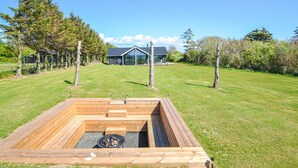 Une terrasse en bois avec un foyer au centre, entourée d’une pelouse bien entretenue et d’arbres.