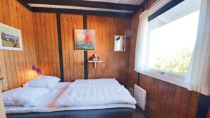 Une pièce aux murs en bois, un lit avec une literie blanche, un tableau accroché au mur, une petite étagère avec quelques objets, et une fenêtre avec des rideaux.
