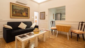 Una sala de estar con un sofá negro, una mesa de centro blanca con revistas, una mesita de madera y un sillón con cojín.