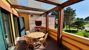 Un balcón techado con una mesa y sillas de madera, una parrilla de ladrillo y vistas a un paisaje verde.