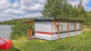 A small, single-story RV-style cabin with a metal roof and wooden accents, situated by a lake with a grassy foreground and trees in the background.