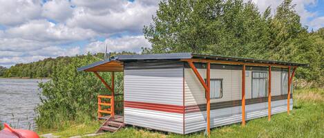 Una cabaña pequeña de un solo piso, al estilo camper, con techo metálico y detalles de madera, ubicada junto a un lago con un primer plano de césped y árboles al fondo.