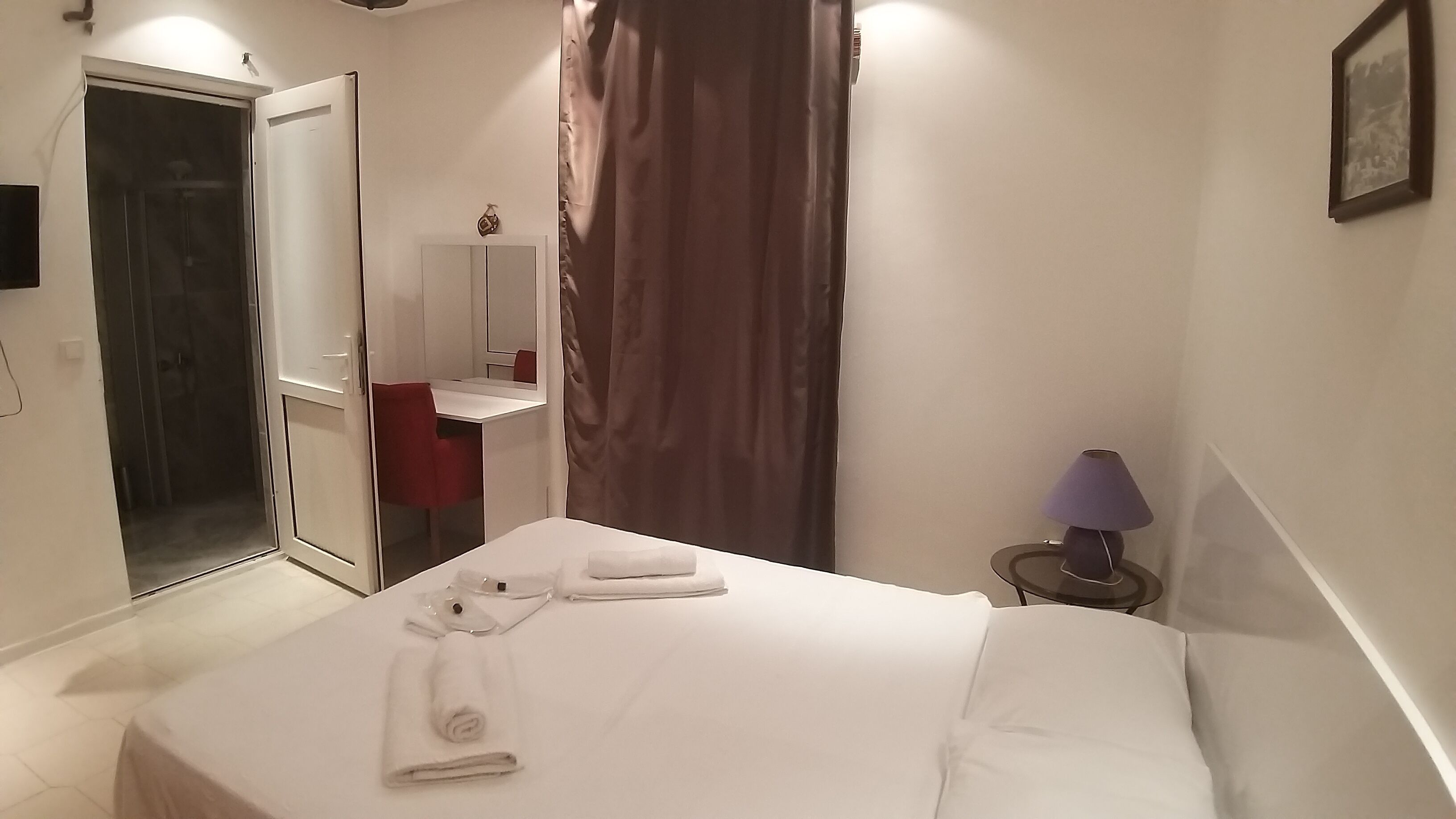 A hotel room with a bed, a desk with a chair, a mirror, a lamp, and a door leading to a bathroom.
