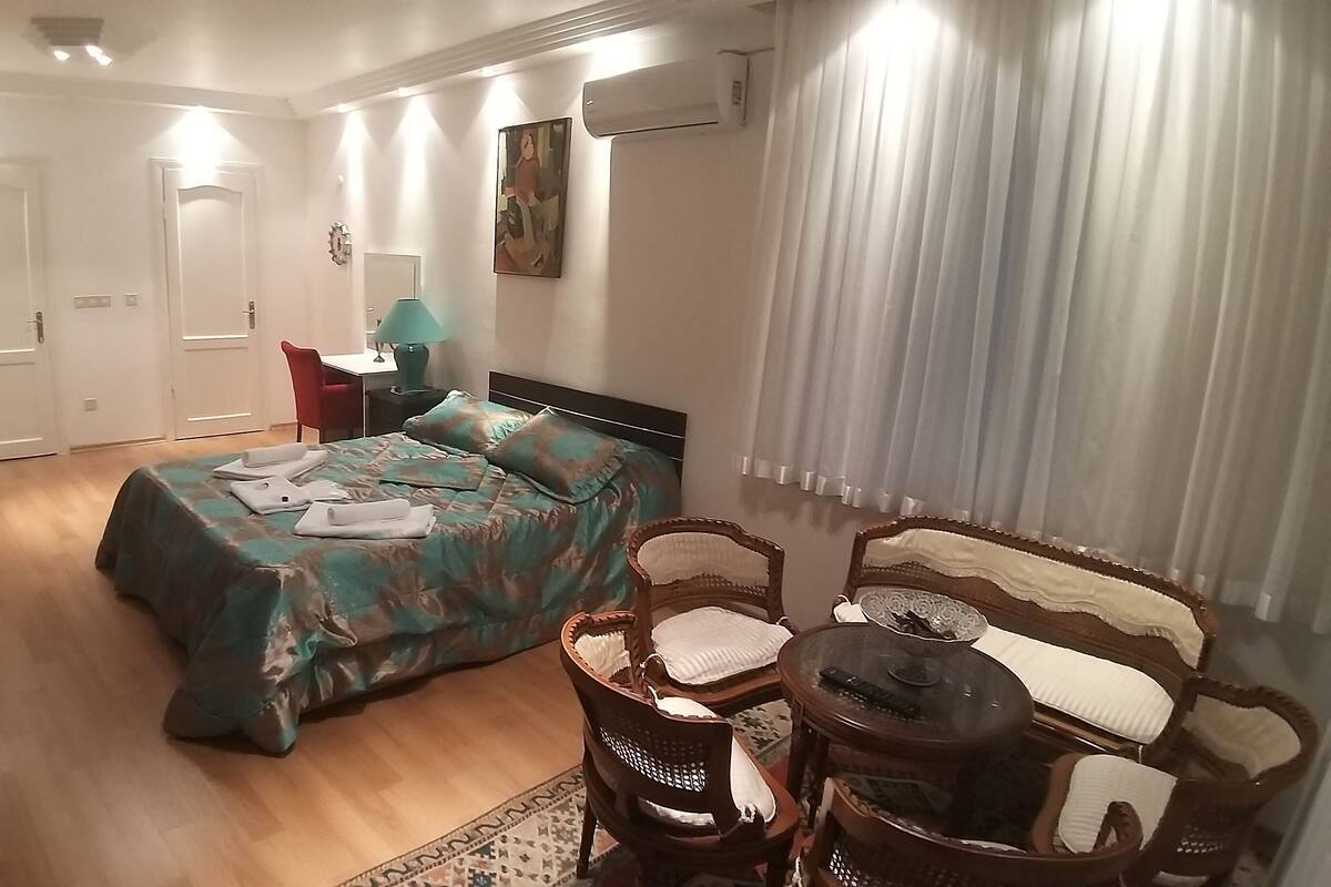 A bedroom with a bed, a chair, a small table, a desk, and a painting on the wall.