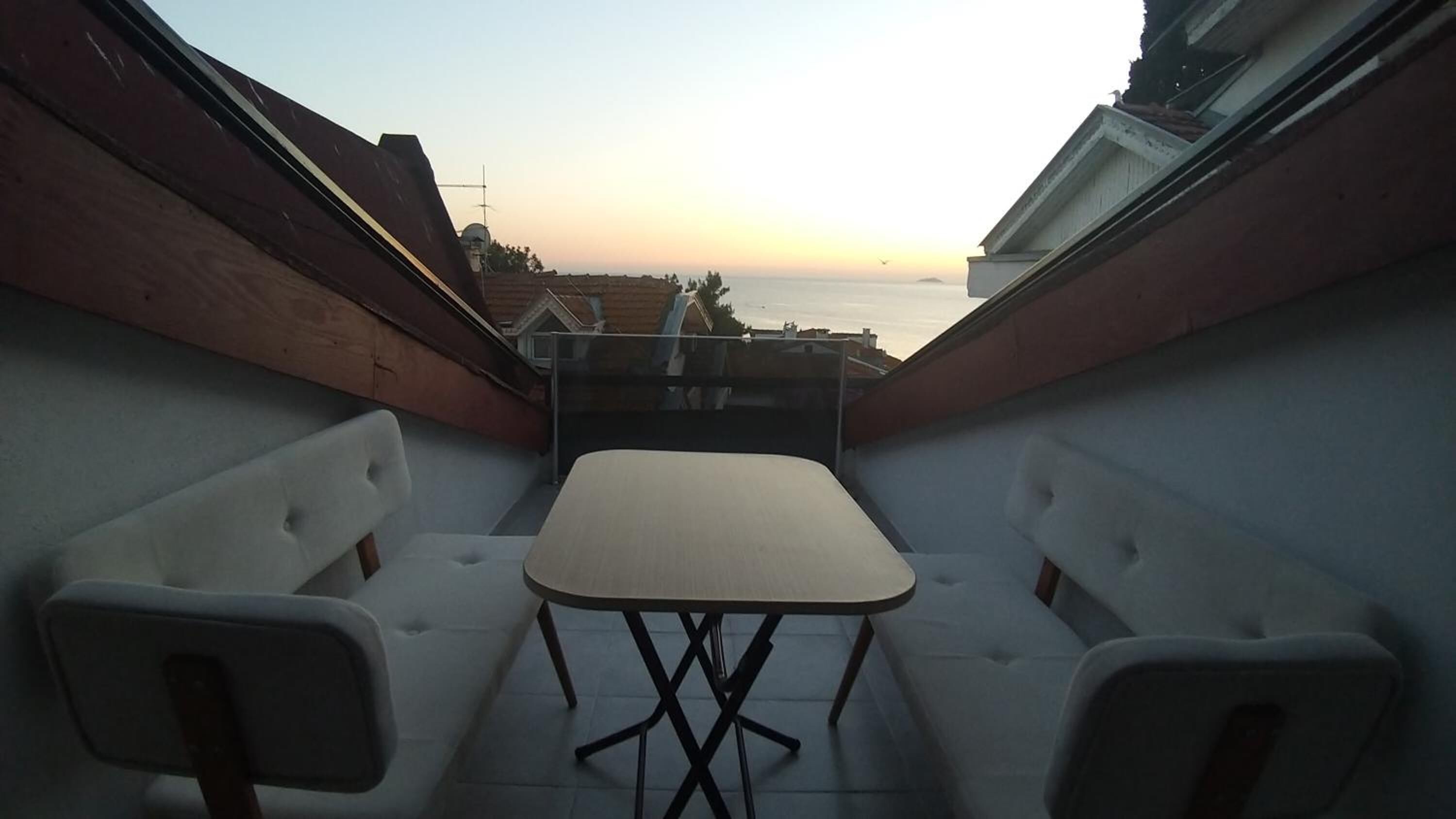 Un pequeño balcón con una mesa y dos sillas, con vistas a una zona residencial al atardecer.