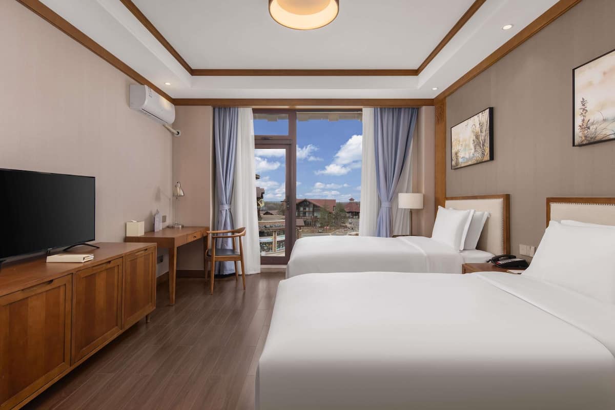 A hotel room with a television, a desk, two beds, a large window with curtains, and a ceiling light.
