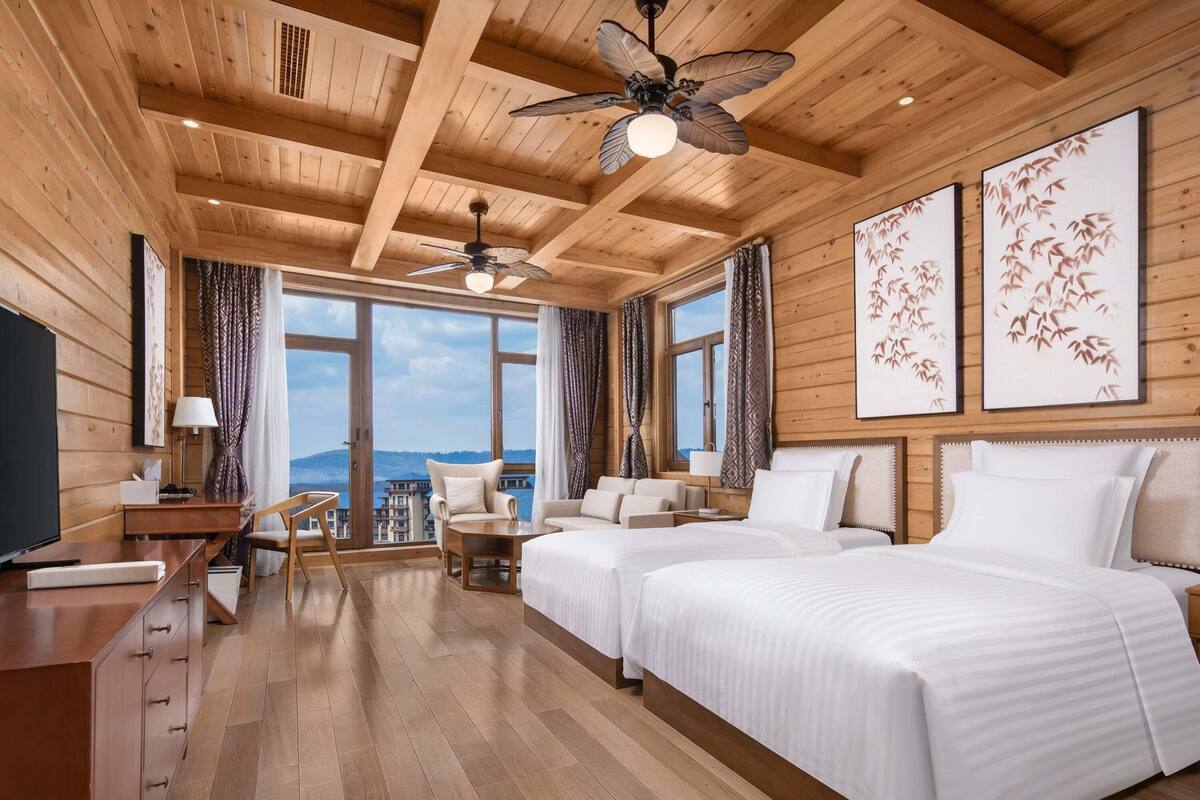 A hotel room with wooden ceilings, two beds, a desk, a chair, and large windows offering a view.