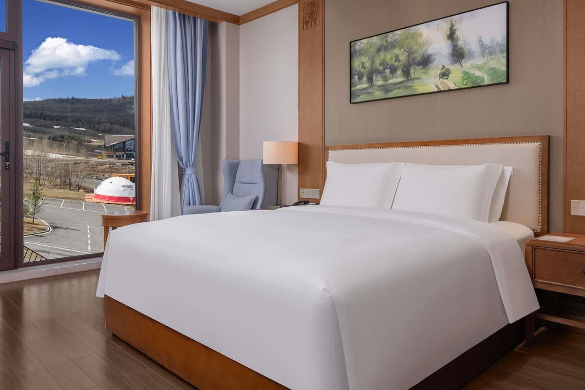 A hotel room with a large bed, a chair, a desk, a lamp, and a painting on the wall.