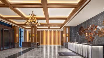 A modern hotel lobby with a high ceiling, a central chandelier, a reception desk, and a decorative wall.