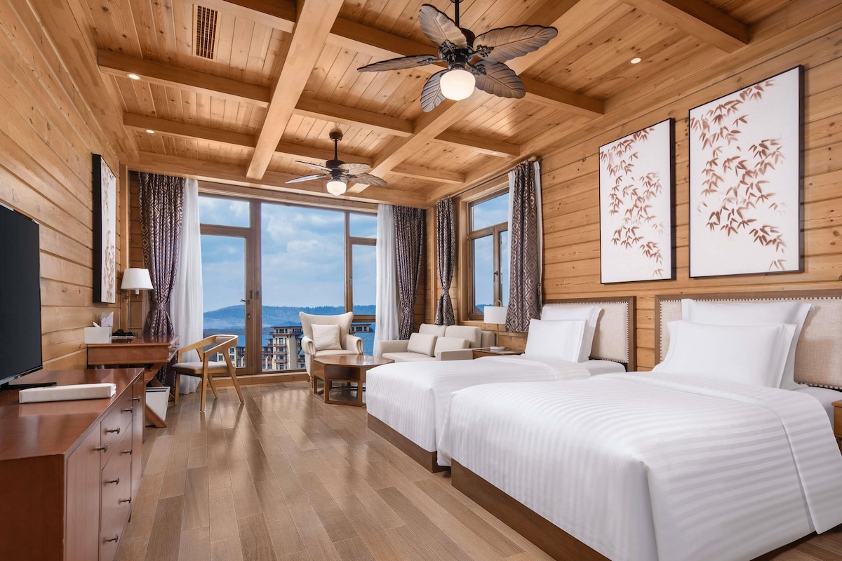 Habitación de hotel con techos de madera, dos camas, un escritorio, una silla y ventanas amplias con vistas.