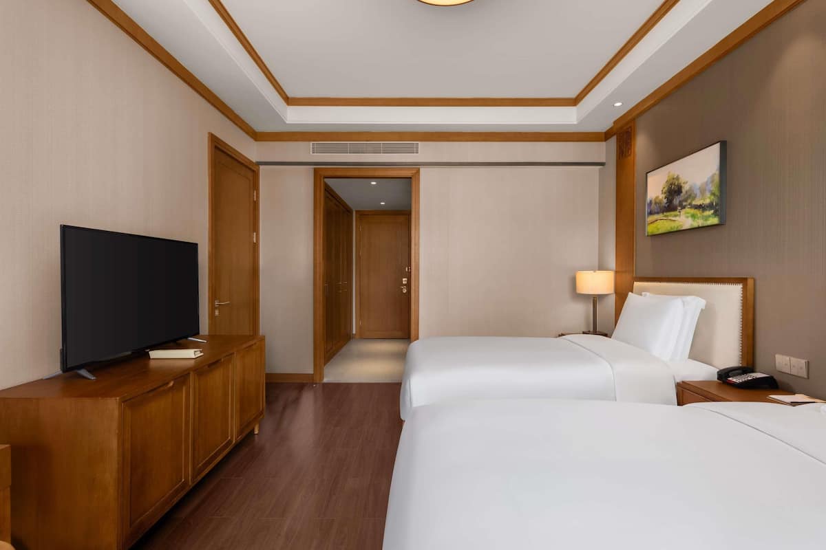 A hotel room with a TV, wooden furniture, a bed, a bedside lamp, and a painting on the wall.