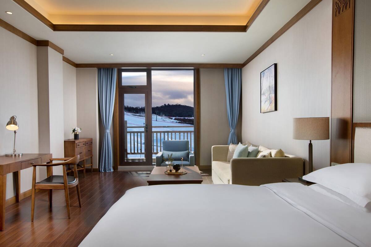 A hotel room with a bed, a desk, a chair, a sofa, and a view of a snowy landscape through a glass door.