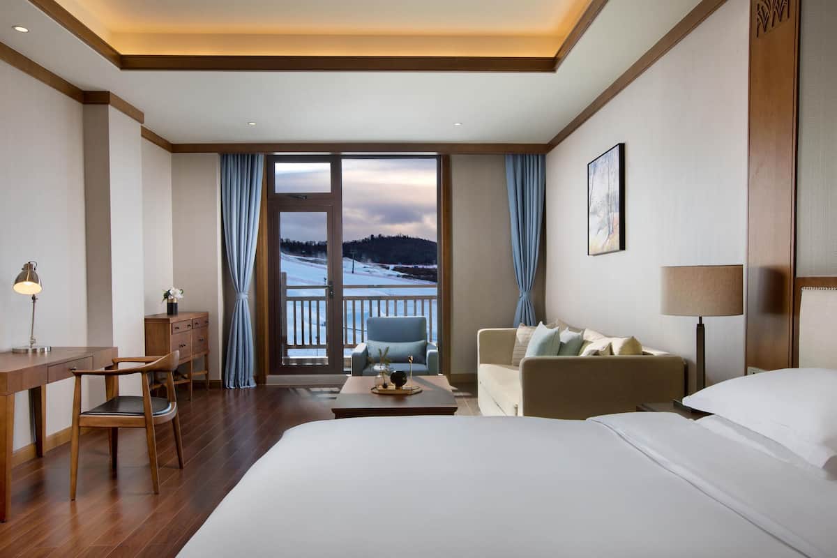 A hotel room with a bed, a desk, a chair, a sofa, and a view of a snowy landscape through a glass door.