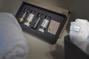 Un ensemble de produits de toilette présenté dans une boîte noire avec une étiquette, posé sur un lit avec une serviette blanche.