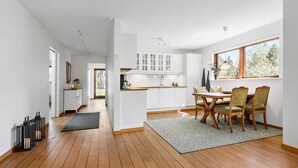 Eine moderne Küche mit weißen Schränken, ein Essbereich mit einem Holztisch und Stühlen sowie ein Blick auf Bäume durch ein Fenster.