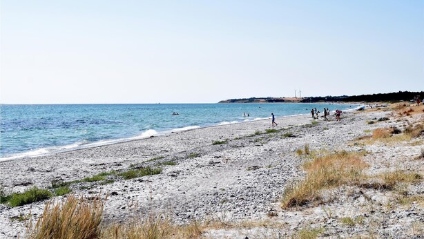 Ein Strand mit Menschen, die am Ufer entlanggehen, einem klaren Himmel und Windkraftanlagen in der Ferne.