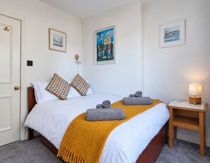 Un lit bien fait, avec une couette blanche, deux coussins à motifs et un plaid jaune. On y voit une table de chevet en bois, une lampe et un tableau encadré accroché au mur.