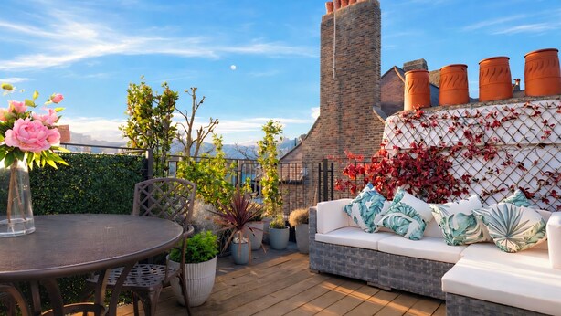 A patio with a round table, chairs, a cushioned seating area, potted plants, and a brick chimney.