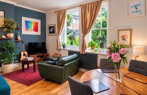 A living room with a green sofa, a wooden coffee table, a TV, and a dining table with chairs. There are potted plants and artwork on the walls. - Beautiful Highgate 2-Bed Apartment | Sunny Roof Terrace (London)