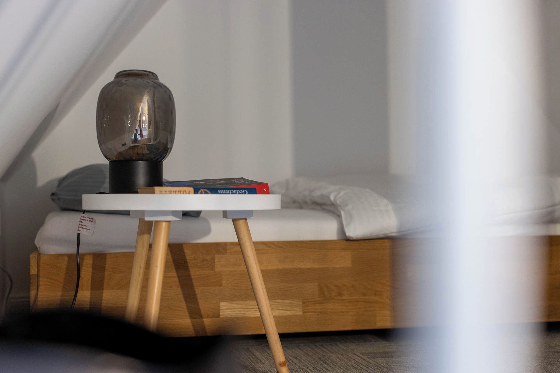 A bedside table with a lamp, books, and a white pillow on a wooden bed frame.