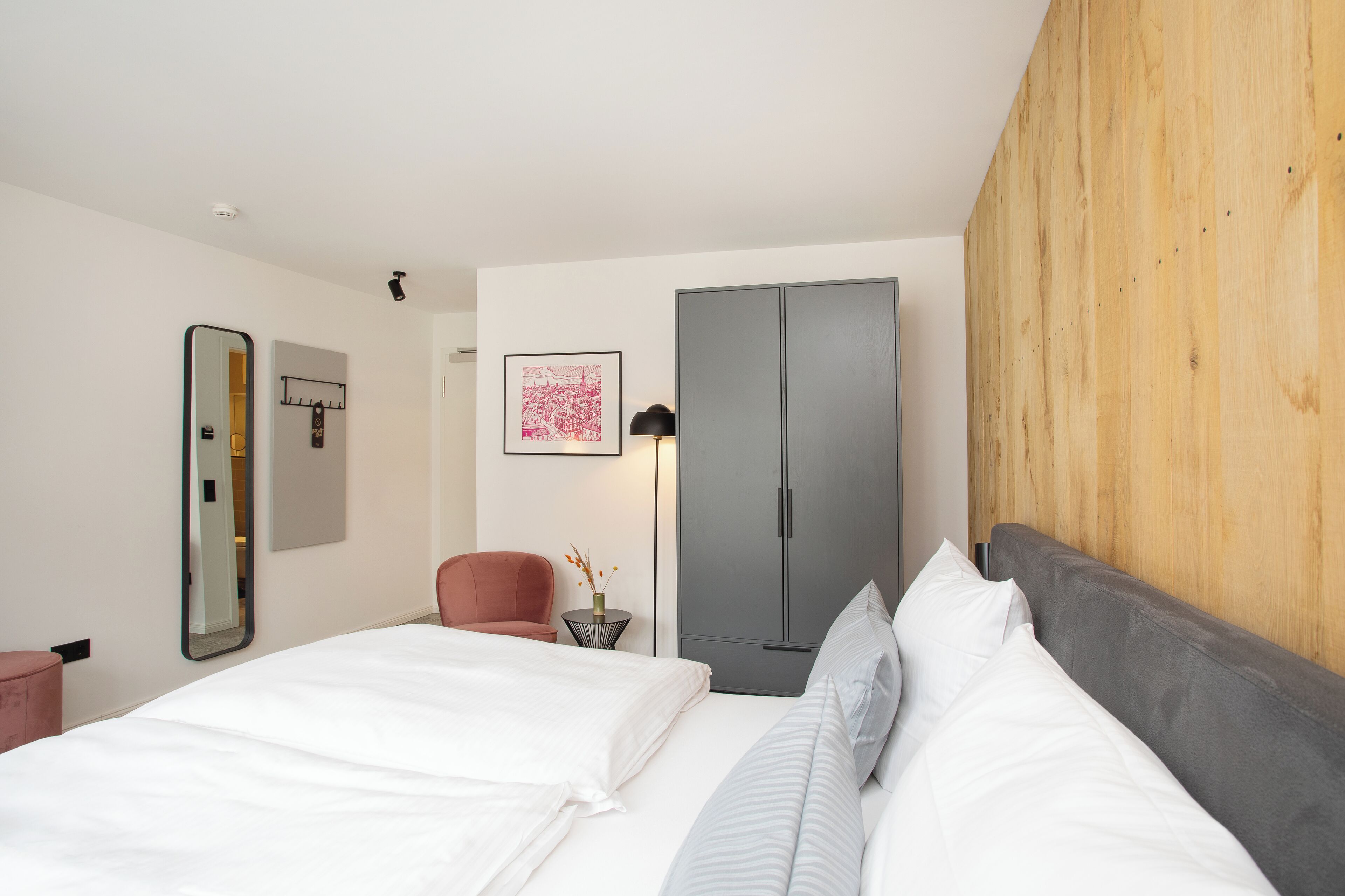 Una habitación de hotel moderna con una cama, un armario, una silla y una mesita de noche con una lámpara.