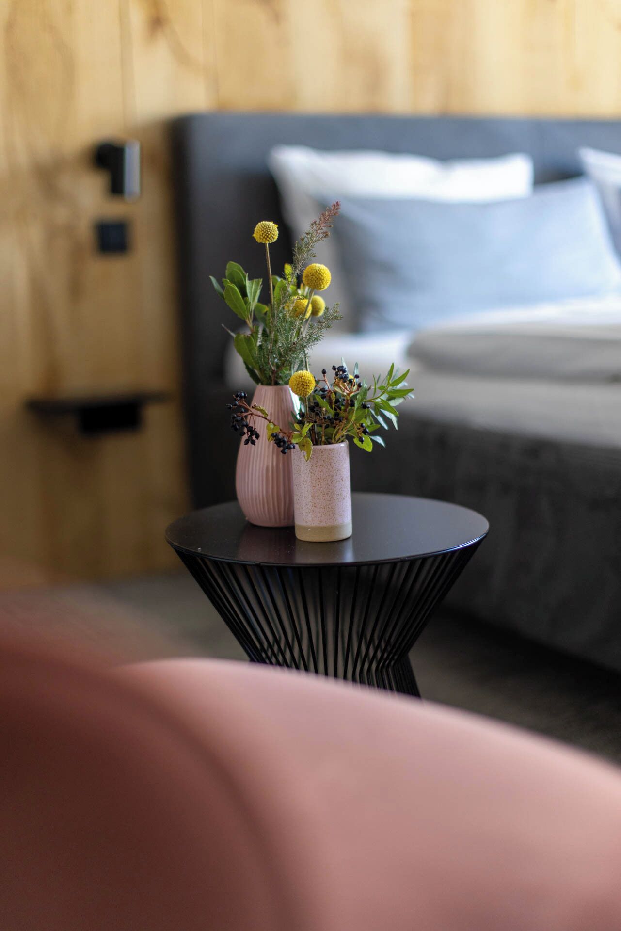 Una mesa redonda pequeña con dos jarrones de plantas en una habitación con una cama y una pared de madera.