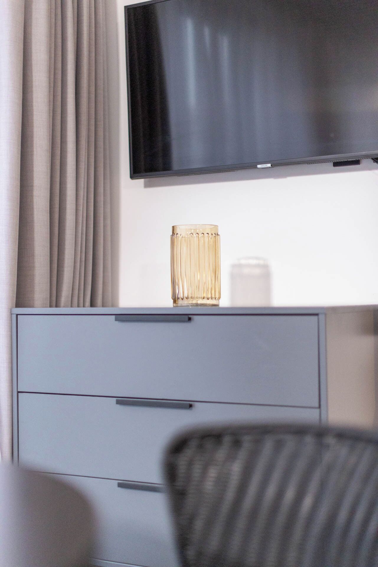 A modern hotel room with a flat-screen TV mounted on the wall, a grey cabinet with drawers, and a striped chair.
