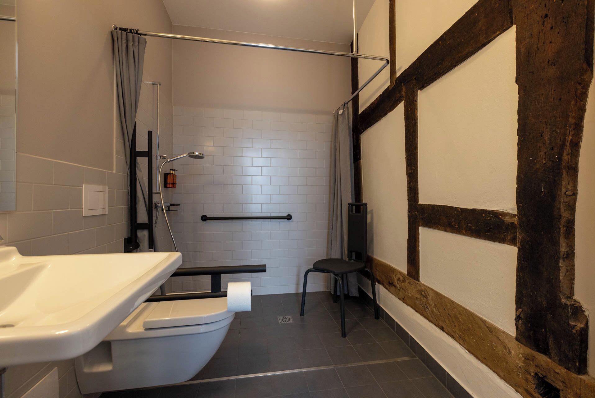 Un baño pequeño con inodoro blanco, ducha con cabezal de ducha de mano y una silla pequeña.