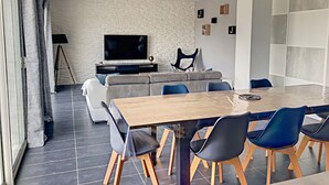 Uma sala de estar moderna com um sofá cinza, uma mesa de jantar de madeira com cadeiras azuis, uma televisão de tela plana em um suporte preto e uma parede de tijolos brancos.