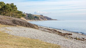 Eine Küstenlandschaft mit Kiesstrand, grünen Wiesen und einem Hügel mit Bäumen und einer Bebauung.