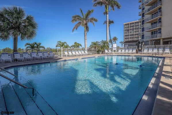 A clear blue pool surrounded by white lounge chairs and palm trees, with a multi-story building in the background.