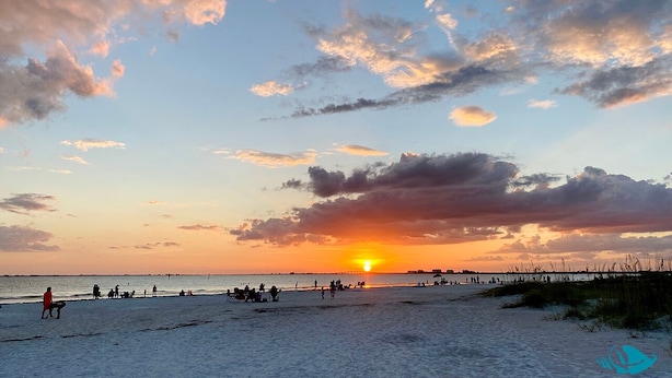 A beach scene with people walking and sitting, a pier in the background, and a sunset. - Pet Friendly Beachfront Vacation Condo At Bargain Price! (Fort Myers Beach)
