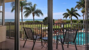 A balcony with two chairs, a pool, and a view of the ocean and palm trees. - Pet Friendly Beachfront Vacation Condo At Bargain Price! (Fort Myers Beach)