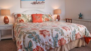 A bedroom with a floral bedspread, two bedside lamps, a beach-themed painting, and a small decorative starfish on the dresser. - Pet Friendly Beachfront Vacation Condo At Bargain Price! (Fort Myers Beach)
